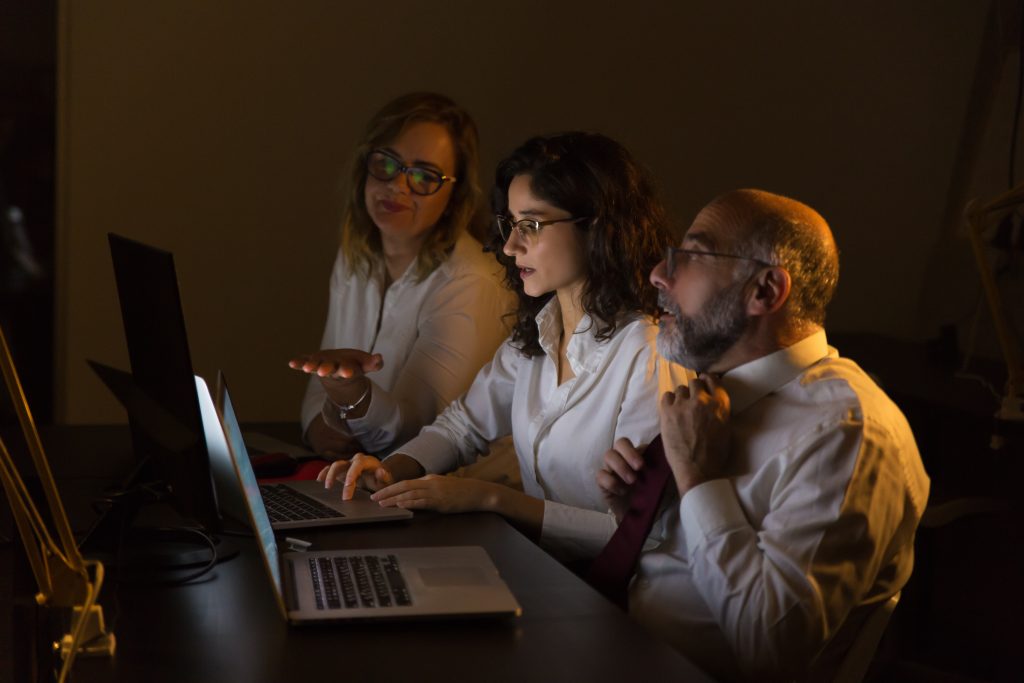 Three professionals in a dimly lit room, huddled around laptops late at night, deep in discussion—capturing the intensity and collaboration behind creative problem-solving.